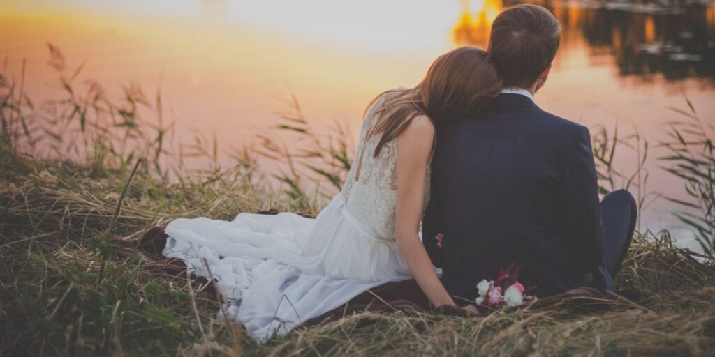 newlywed couple sitting by the water while bride has her head on groom's shoulder